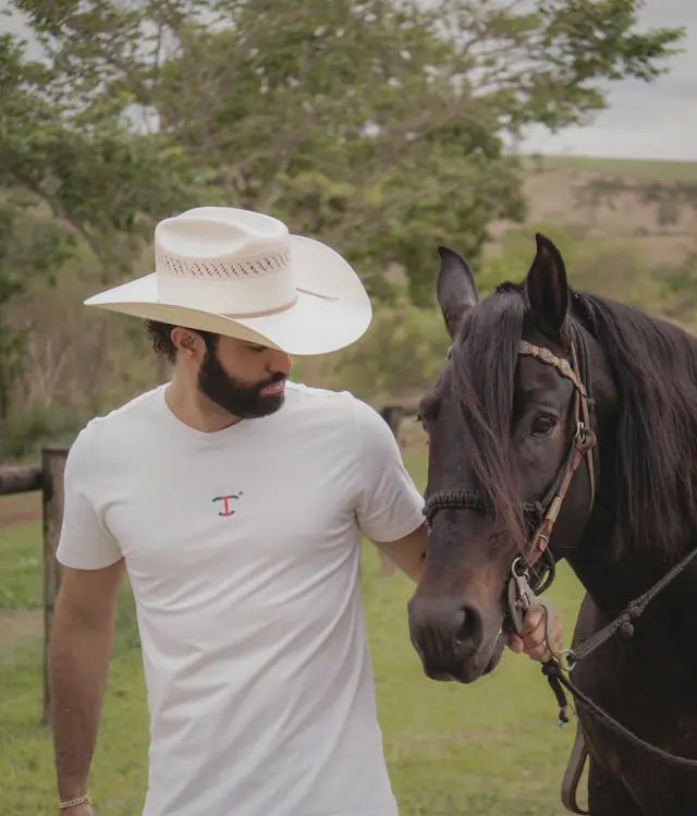 Homem de chapéu de palha e camiseta clara com o monograma T da TBull bordado no peito, de pé ao lado de um cavalo preto, em pasto com cerca de madeira ao fundo.
